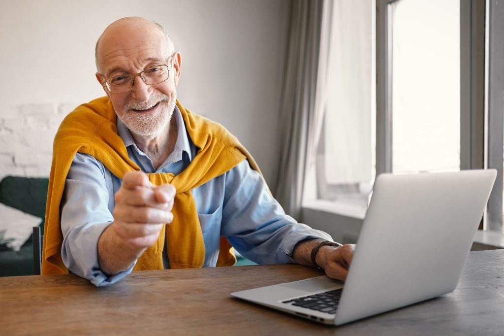 fashionable friendly elderly male recruiter wearing rectangular glasses elegant clothes working portable computer smiling broadly pointing finger choosing you job position (1)